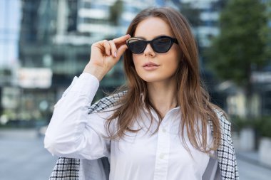 Kendine güveni tam bir iş kadını şık giyinmiş şehir merkezinde cam binalarla dikiliyor. Modern şehir kadını şık kıyafetler ve güneş gözlükleriyle kurumsal gökdelenlerin önünde poz veriyor..