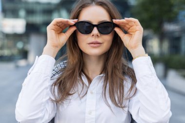 Şehir sokağında kendine güvenen ve tarz sahibi genç bir profesyonel şık ofis kıyafetleri giyiyor. Şık bir kadın, şehir ortamında güven içinde poz vererek güneş gözlüğü takıyor. Modern iş