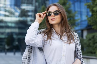 Finansal bölgede güneş gözlüğü ve şık kıyafetler takan zarif genç bir profesyonel. Güneş gözlüklü şık bir kadın şehir ortamında güven içinde poz veriyor. Modern iş kıyafeti ve c