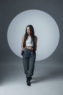 Kendine güveni tam, beyaz bluzlu, kot pantolonlu ve botlu dramatik ışıklandırması ve dairesel bir sahne ışığı olan bir stüdyoda poz veren genç bir kadın. Moda, tutum ve tarz kavramı.