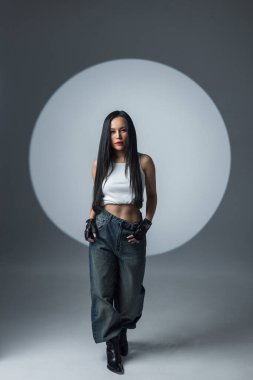 Ekin bluzlu, kot pantolonlu, botlu havalı bir kadın modern bir stüdyoda dairesel bir spot ışığı altında yürüyor..