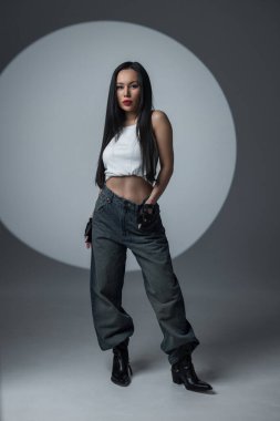 Uzun saçlı, kendine güvenen bir manken sokak giysisi tarzında cesur, yuvarlak bir arka planın önünde yürüyor..