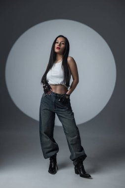 Kendine güveni tam, beyaz bluzlu, kot pantolonlu ve botlu dramatik ışıklandırması ve dairesel bir sahne ışığı olan bir stüdyoda poz veren genç bir kadın. Moda, tutum ve tarz kavramı.