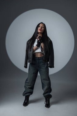 Deri ceketli şık bir kadın, etekli bluz ve bol kotlu dramatik ışıklandırmayla stüdyoda güven içinde poz veriyor. Moda modeli cesur makyajlı ve kenar mahalle tarzında..