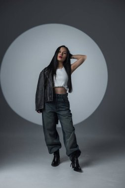 Modern bir stüdyoda duran siyah deri ceketli ve yüksek bel kotlu şık bir bayan model. Güçlü, şehirli ve kendine güvenen moda konsepti. Asyalı güzel kadın