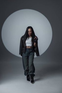 Kenar mahalle kıyafetleri içindeki kendine güvenen kadın dramatik bir sahne ışığı altında poz veriyor. Deri ceket, bol kot pantolon ve eldiven giyerek güç ve tavır sergiliyor..