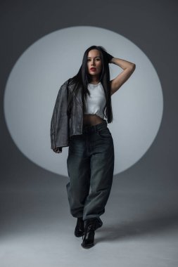 Uzun siyah saçlı, şık bir kadın stüdyo ışıklarında şiddetle poz veriyor. Sokak tarzı kıyafetler ve koyu kırmızı ruj sürerek kendine güveni ve isyanı simgeliyor..