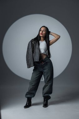 Grunge 'dan esinlenilmiş kıyafetli modern kadın dramatik ışıklandırma altında güçlü bir poz veriyor. Şık deri ceket ve bol kot onun cesur kişiliğini ön plana çıkarıyor..