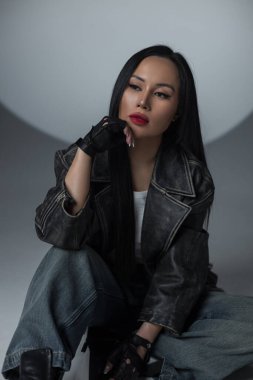 Geniş paçalı kot pantolon ve deri ceketle yerde oturan havalı bir kadın manken. Grunge stili sahne ışığı arka planında stüdyonun cazibesiyle buluşuyor.