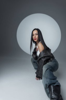 Kenar mahalleli genç bir kadın model yerde poz veriyor. Deri ceket, geniş kot ve stüdyo ışıkları altında botlar güçlü, şık bir hava yaratır..