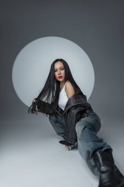 Deri ceketli, beyaz kolsuz tişörtlü ve stüdyo zemininde kendine güvenen kotlu şık genç bir kadın. Göze çarpan arka planı ve dramatik havası olan cesur, gergin bir moda..