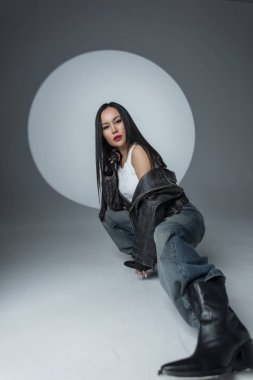 Sokak tarzı giyinmiş kendine güvenen bir kadın stüdyo zemininde oturuyor ve arkasında bir spot ışığı var. Modern moda, deri ceket, kot pantolon ve sert tavır. Asyalı güzel kadın