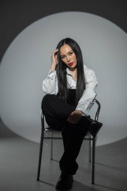Kırmızı dudaklı, şık giyinmiş, beyaz gömlekli, siyah korseli güzel bir Asyalı kadın fotoğraf stüdyosunun arka planında, koyu renk koltukta oturuyor. Güzel kız.