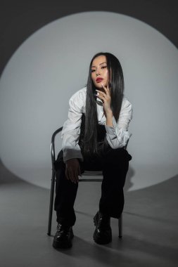 Kırmızı dudaklı, şık giyinmiş, beyaz gömlekli, siyah korseli güzel bir Asyalı kadın fotoğraf stüdyosunun arka planında, koyu renk koltukta oturuyor. Güzel kız.