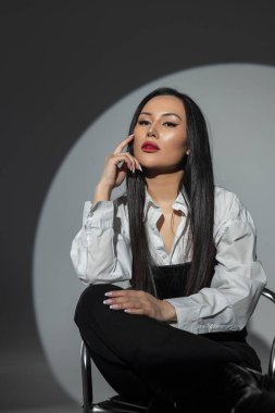 Kırmızı dudaklı, şık giyinmiş, beyaz gömlekli, siyah korseli güzel bir Asyalı kadın fotoğraf stüdyosunun arka planında, koyu renk koltukta oturuyor. Güzel kız.