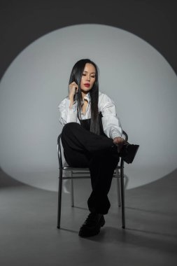 Kırmızı dudaklı, şık giyinmiş, beyaz gömlekli, siyah korseli güzel bir Asyalı kadın fotoğraf stüdyosunun arka planında, koyu renk koltukta oturuyor. Güzel kız.