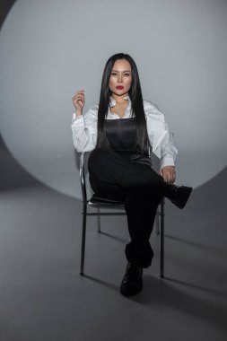 Uzun siyah saçlı, stüdyo ışıklarında poz veren kendine güvenen bir kadın. Tek renkli kıyafet, koyu kırmızı dudaklar, minimum arkaplan. Güçlü, şık ve profesyonel bir portre..