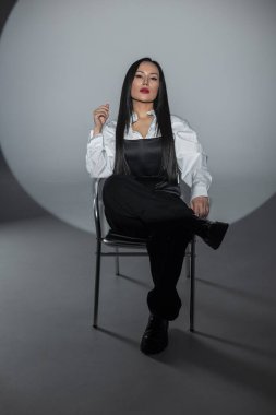 Şık manken spot ışıkları altında sandalyede oturur. Beyaz gömlek ve siyah pantolon giyerek güç ve entelektüellik yayıyor. Dramatik stüdyo ortamında yüksek moda editör havası.