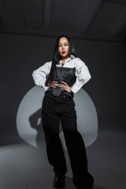 Siyahlar ve beyazlar içindeki kendine güvenen Asyalı kadın dramatik bir sahne ışığı altında poz veriyor. Cesur kırmızı dudaklar, güçlü duruş, modern tarz. Stüdyo portresi, güçlü tavır ve asgari arkaplan.