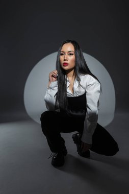 Karanlık stüdyo ortamında, cesur moda giyinen ve dramatik ışıklandırma altında kendine güvenen genç şık bir kadın. Minimalist, kentsel estetik.