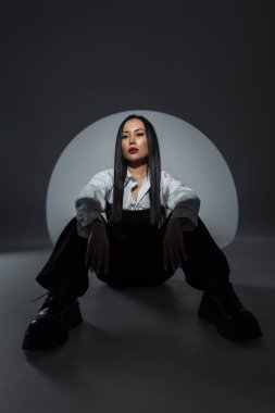 Asyalı güzel kadın. Beyaz gömlekli ve siyah pantolonlu manken stüdyo zemininde dramatik ışıklandırmayla oturuyor. Cesur makyaj ve ciddi ifade. Şık kentsel görünüş.
