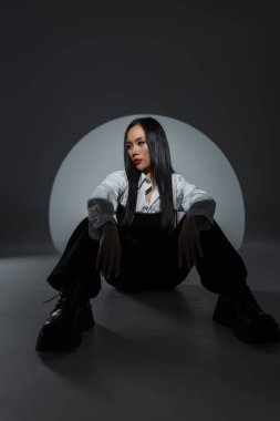 Modayı takip eden kadın model dairesel bir ışığın altında tek renkli kıyafetle yerde oturuyor. Minimalist tarz, karamsar atmosfer. Asyalı güzel kadın.