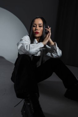 Asyalı güzel kadın. Karanlık stüdyoda kendine güvenen genç bir kadın sahne ışıklarıyla poz veriyor. Beyaz gömlek ve asker çizmeleri giyerek, güç ve modern tavır sergiliyor..