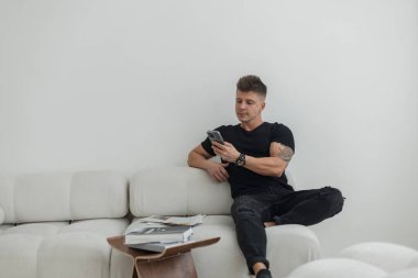 Siyah elbiseli havalı, başarılı yakışıklı adam elinde akıllı telefon haberleri tutarak kanepede oturuyor.