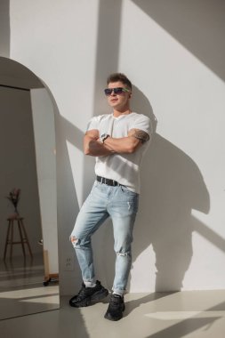 Güneş gözlüklü şık bir adamın portresi, evde öylesine poz veriyor. Beyaz tişört ve açık renk kot giymiş, kolunda dövmesi olan cesur ve modern bir görünüm yakalıyor..