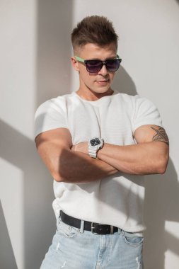 Güneş gözlüğü takan ve evde poz veren kendinden emin bir adam. Parlak güneş ışığı beyaz duvarda dramatik gölgeler yaratır. Kentsel, modern ve şık bir ortam..