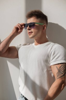 Güneş gözlüklü şık genç adam rahat bir şekilde beyaz tişört ve yırtık kotla poz veriyor. Modern, kapalı alan ayarları ve doğal güneş ışığı.