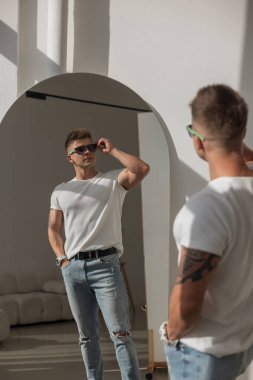 Beyaz tişörtlü ve kotlu dövmeli adam aynadaki yansımasına bakıyor. Parlak modern iç mekan, güneş ışığı gölge düşürüyor. Kendine güvenen ve şık bir hava..