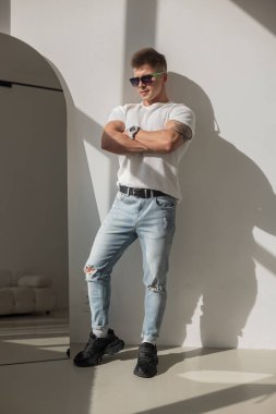 Gündelik kıyafetli ve güneş gözlüklü kendinden emin genç adam parlak güneş ışığında poz veriyor. Beyaz gömlek, yırtık kot pantolon, siyah spor ayakkabı ve kolları çapraz duran bir saat..