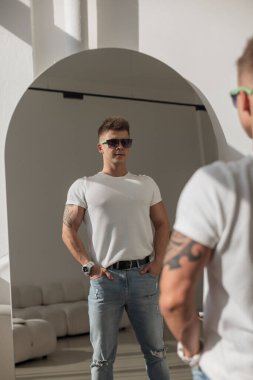 Dövmeli ve güneş gözlüklü şık bir genç adam aynada kendine bakıyor. Beyaz bir t-shirt ve yırtık kot pantolon giyiyor. Minimalist dekorlu, güneşli, modern bir iç mekan..