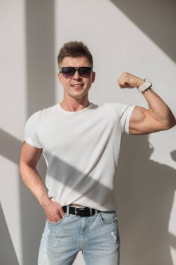 Güneş gözlüklü, pazılarını esneten, beyaz tişört ve yırtık kot giyen, gün ışığında keskin gölgeli beyaz bir duvara karşı duran kendine güvenen genç bir adam. Sıradan ve sportif görünüm.