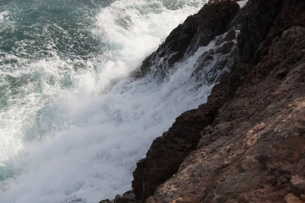 Deniz dalgaları kayalık kıyılara çarpıyor, su sıçrıyor.