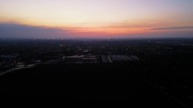 Gökyüzüne turuncu, pembe ve mor renkler yayılan canlı bir günbatımı giderek daha karanlık bir ufukta kayboluyor. Aşağıda, şehir gölgelerle kaplanmış, küçük bir ışık parıltısı kentsel aktiviteyi gösteriyor..