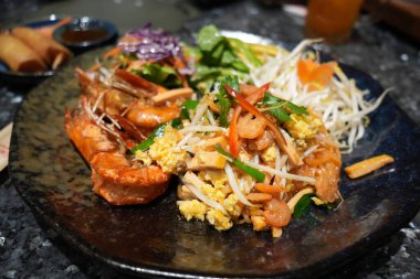 Pad Thai kahverengi ahşap bir masaya yerleştirilir. Daha fazla baharat eklemek için limon suyu sıkan bir kadın var. Çünkü Taylandlılar baharatlı yemek yemeyi sever. Ünlü Tayland yemekleri konsepti. Sokak yemeği. Tayland kimliği