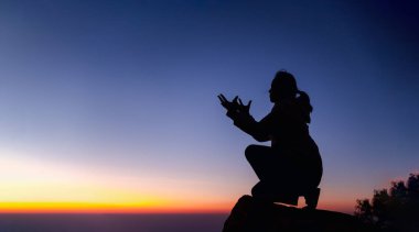 Silhouette of woman kneeling down praying for worship God at sky background. Christians pray to jesus christ for calmness. In morning people got to a quiet place and prayed. Banner with copy space.
