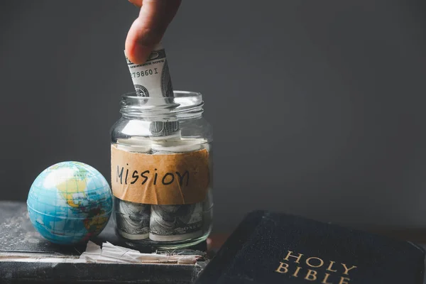 Saving jars full of money and globe with Holy Bible for mission, Mission christian idea. Hand holding dollar with bible on wooden table, Christian background for great commission or earth day concept.