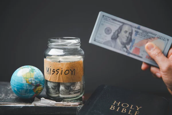 Saving jars full of money and globe with Holy Bible for mission, Mission christian idea. Hand holding dollar with bible on wooden table, Christian background for great commission or earth day concept.