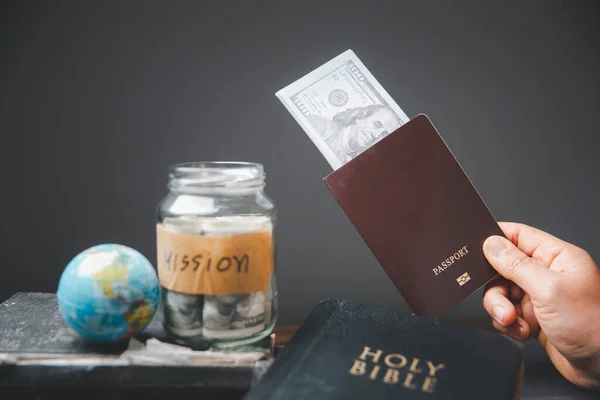 Saving jars full of money and globe with Holy Bible for mission, Mission christian idea. Hand holding dollar and passport on table, Christian background for great commission or earth day concept.