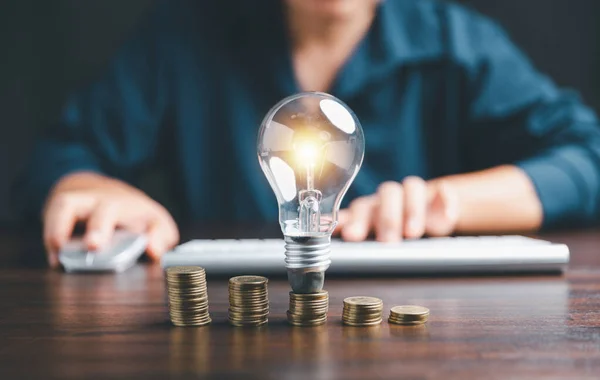 Businesswoman with lightbulb on coins and using calculator to calculate and money stack. Save energy and money with accounting finance in office concept. Idea of energy saving planning in home.