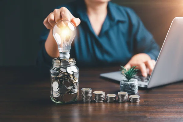 Businesswoman with lightbulb on money jar and using calculator to calculate and money stack. Save energy and money with accounting finance in office concept. Idea of energy saving planning in home.