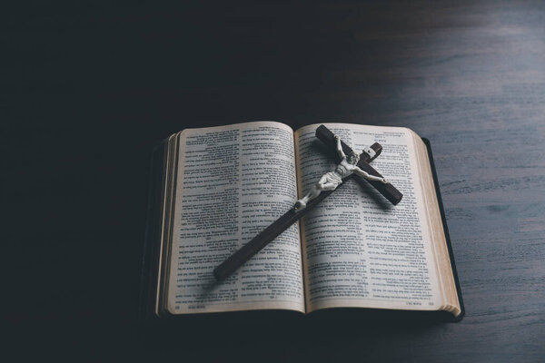Wooden Christian cross crucifix on open holy bible background. Wooden Christian cross on grey table against blurred lights, space for text.