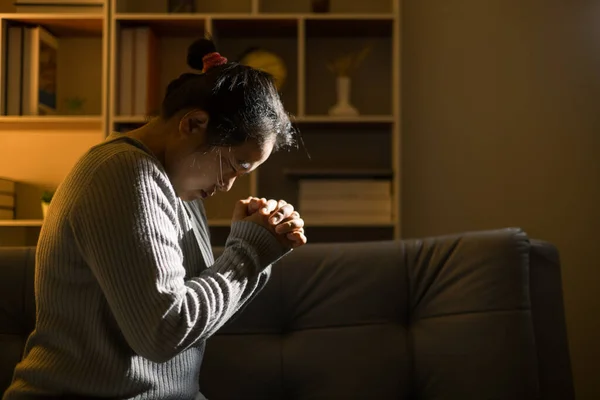Christian religious woman praying at night sitting on couch in living dark room at home. Human person hands worship, Concept of praying to God. Female prayer with holy bible. Lady thanksgiving night