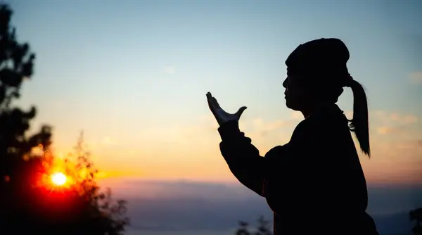 Silhouette of christian woman prayer on sunset background. Woman raising his hands in worship for peaceful life. Female praising God on the mountain. Christian religion concept.