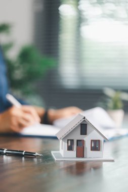 Emlakçı ve müşteri ev satın alma sözleşmesini tartışırken, arka planda emlak, sigorta ve kredi düzenlemelerini sembolize eden ev modeli süreçte yer alıyor.