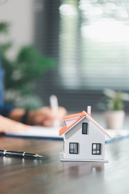 Emlakçı ve müşteri ev satın alma sözleşmesini tartışırken, arka planda emlak, sigorta ve kredi düzenlemelerini sembolize eden ev modeli süreçte yer alıyor.