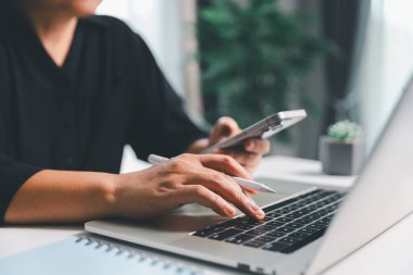 Yakın çekim sahnesinde, masada oturan bir kadın akıllı bir telefon tutuyor, modern e-randevu hizmetleri ve sosyal medyada mobil bir uygulama ile vakit geçiriyor, dijital kullanım kablosuz kavramını gösteriyor.